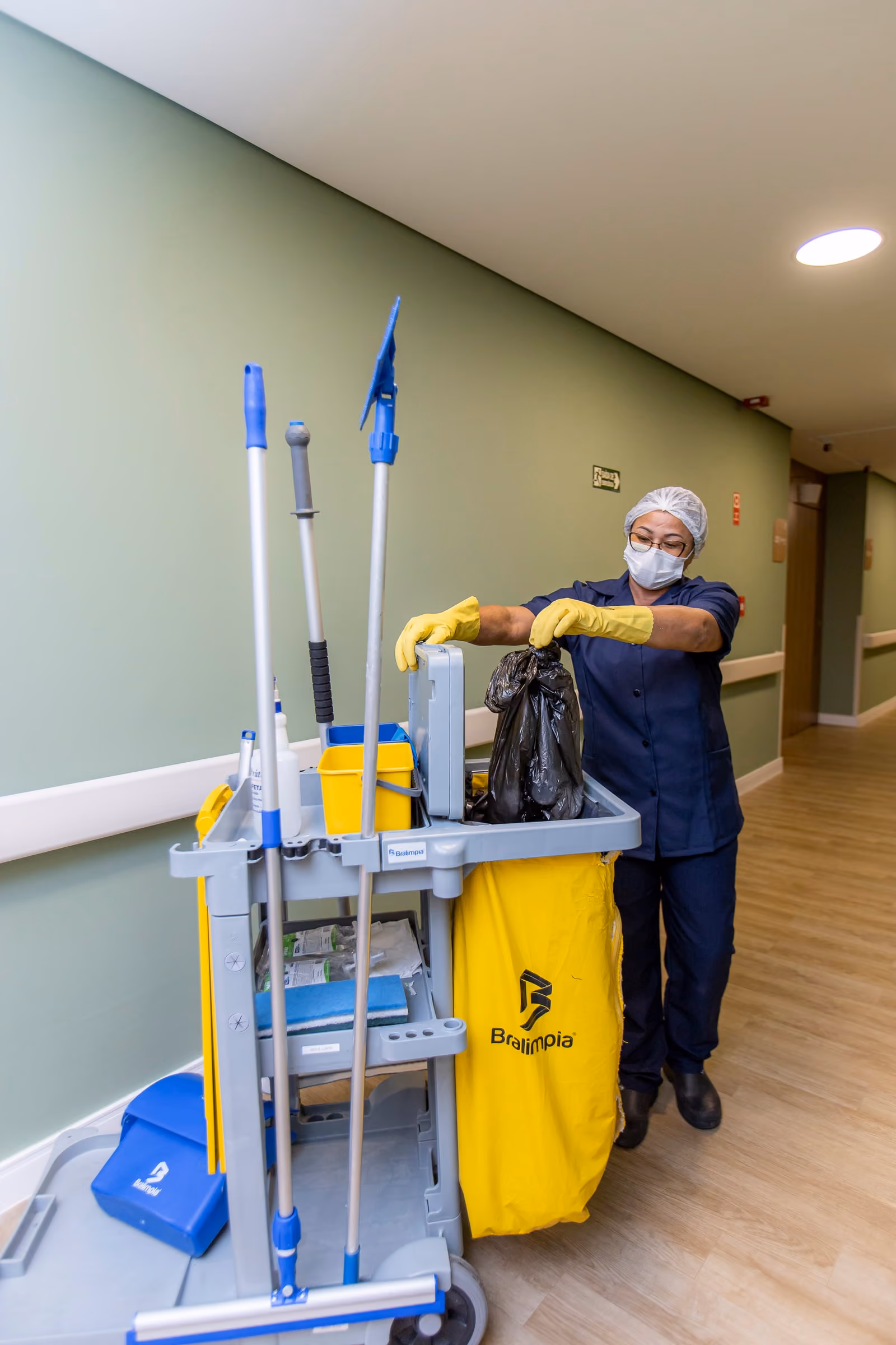 Profissional da Prátika de touca, máscara e luvas recolhe o saco de resíduos no carrinho de limpeza, num corredor hospitalar