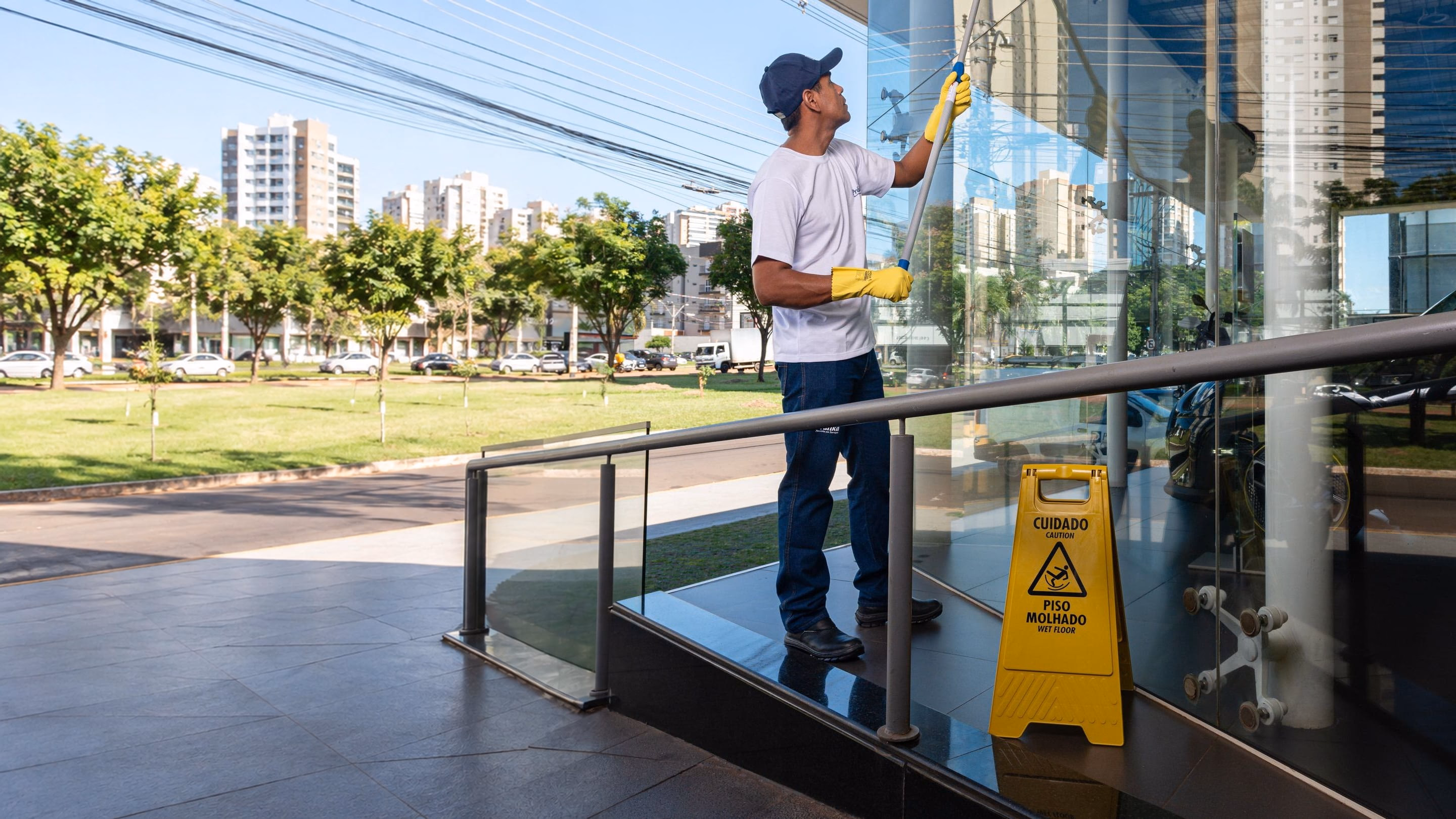 Profissional da Prátika limpa a fachada de vidro de um edifício comercial com rodo de haste longa, placa de piso molhado ao lado