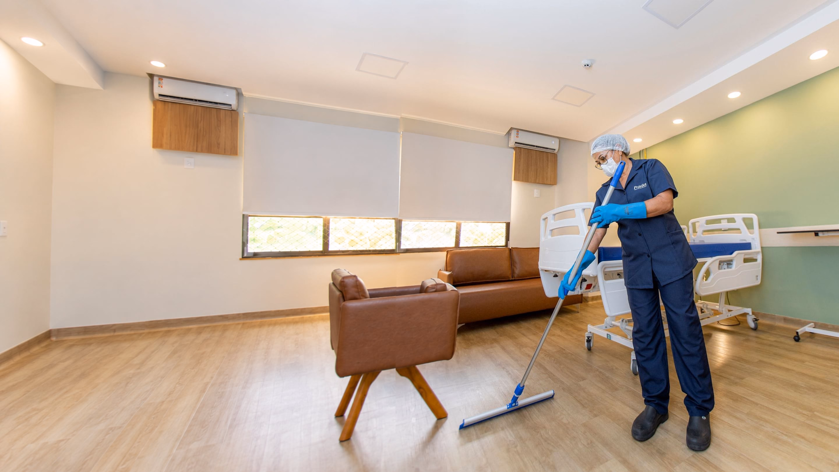 Profissional da Prátika de touca, máscara e luvas passa o rodo com pano no piso de um quarto hospitalar