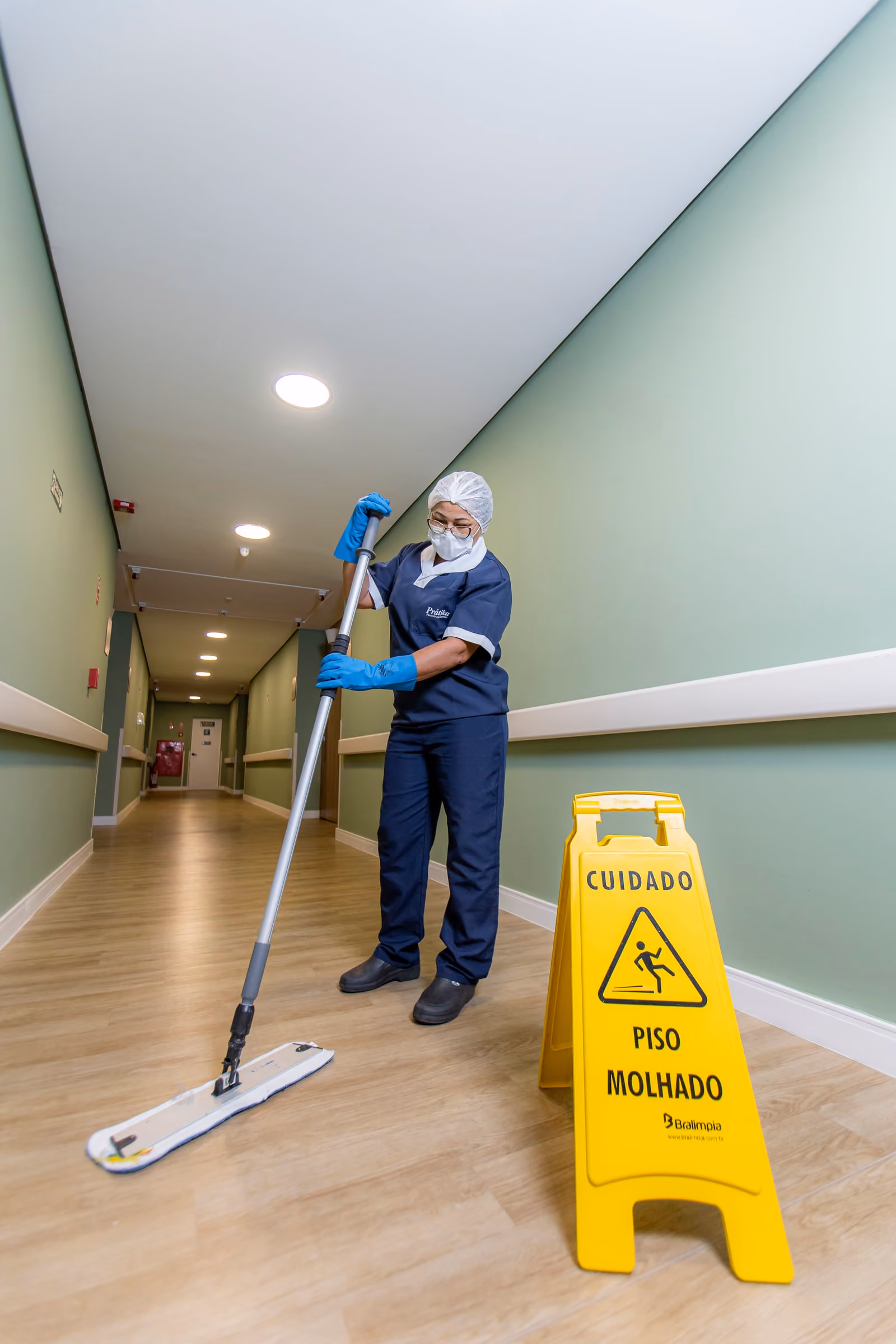 Profissional da Prátika limpa o piso de um corredor hospitalar, com a placa de piso molhado ao lado